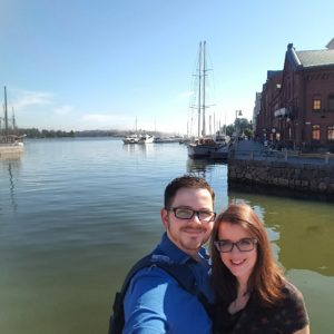 Emma and Dave in Helsinki, standing at a harbour. The water is flat, clear and blue and there are many boats in the background.